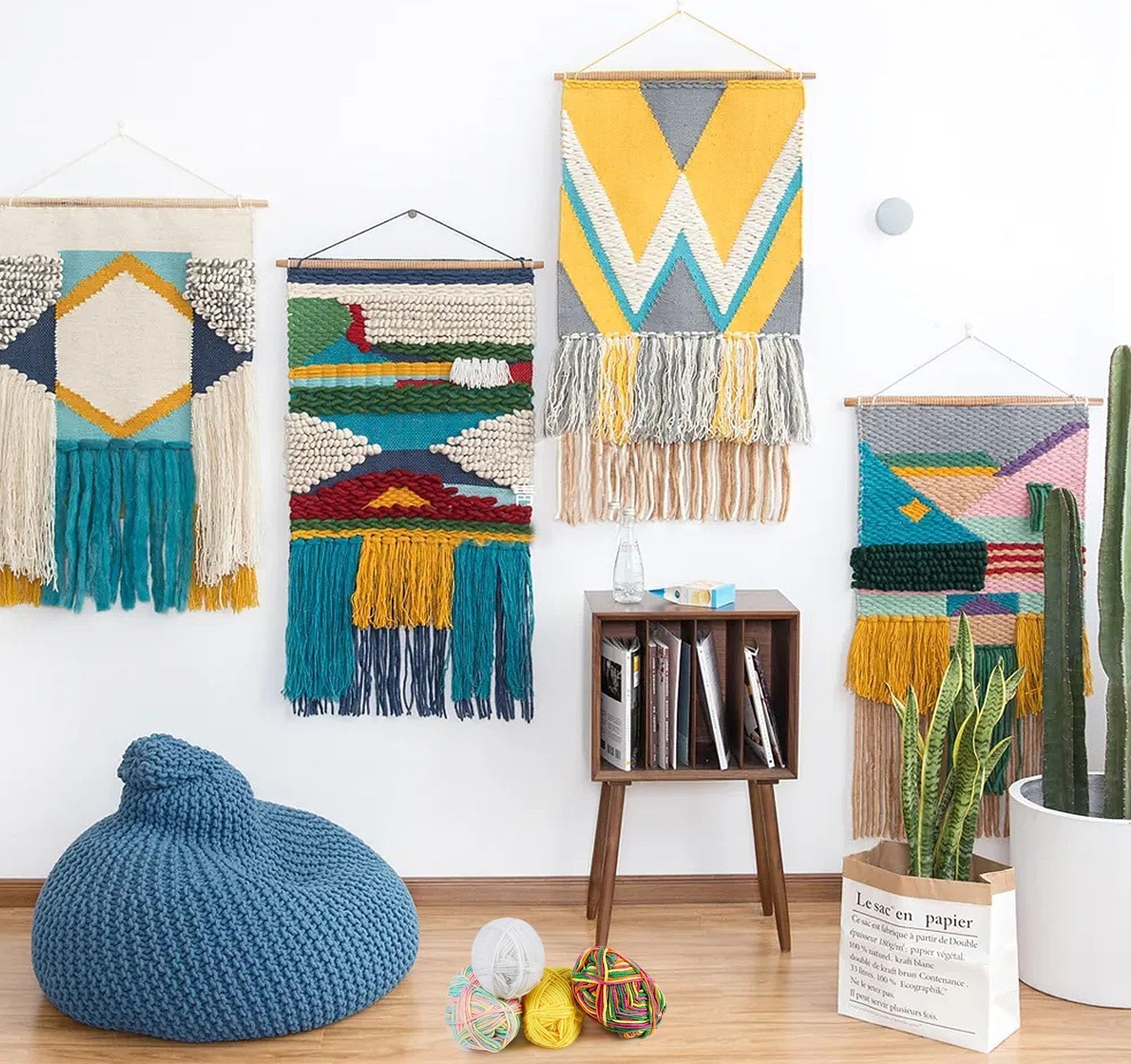 Four woven wall hangings displayed with a blue beanbag chair, yarn balls, a wooden side table with books, and potted plants.