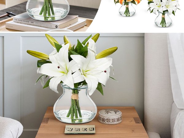 White lily artificial flower arrangement in a clear glass vase with faux water on a wooden nightstand.