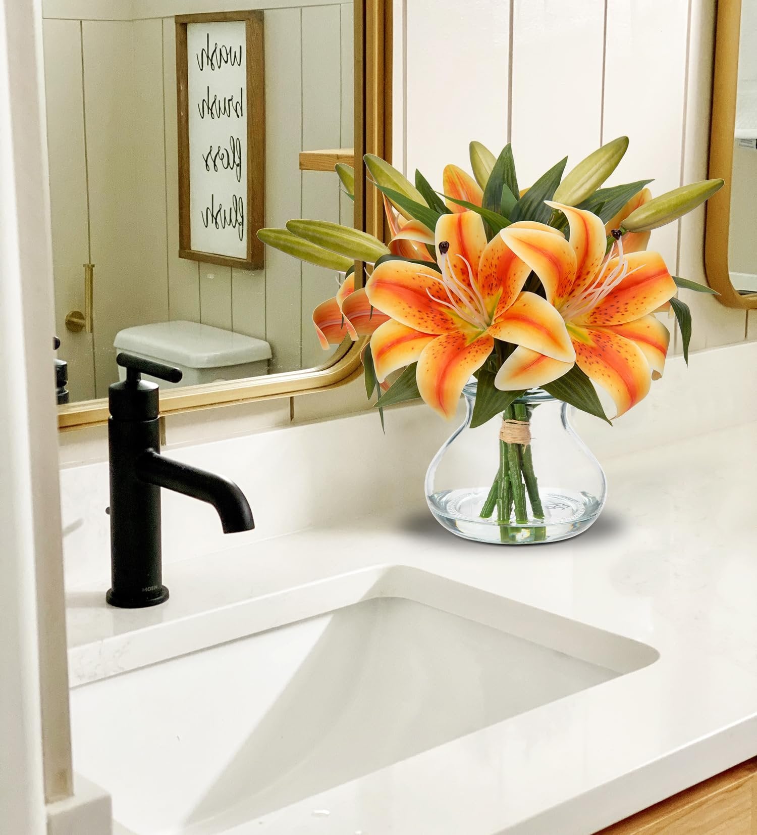Orange tiger lily artificial flower arrangement in a glass vase with faux water on a bathroom counter.