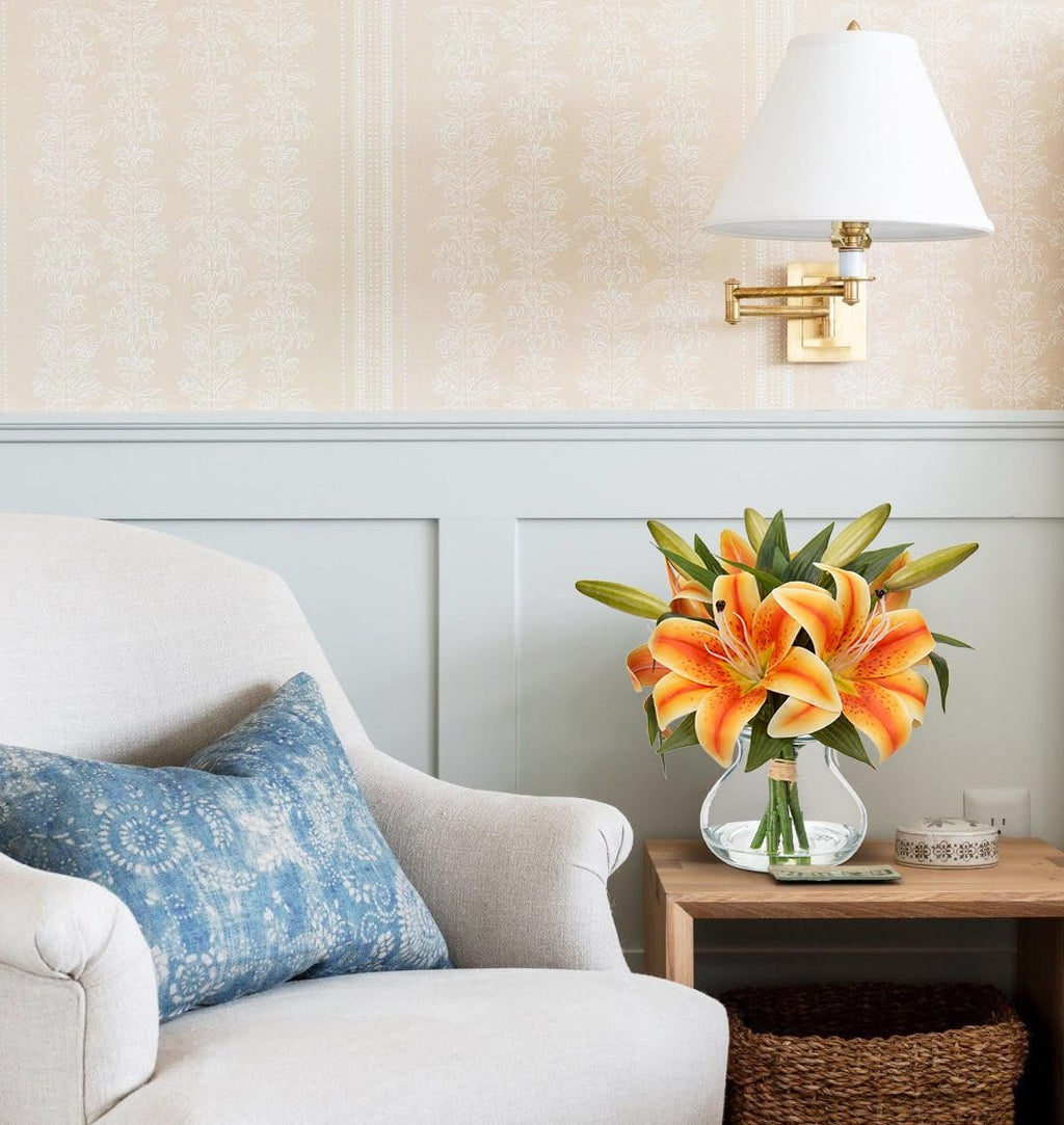 Artificial orange tiger lily arrangement in a glass vase with faux water on a side table next to an armchair.