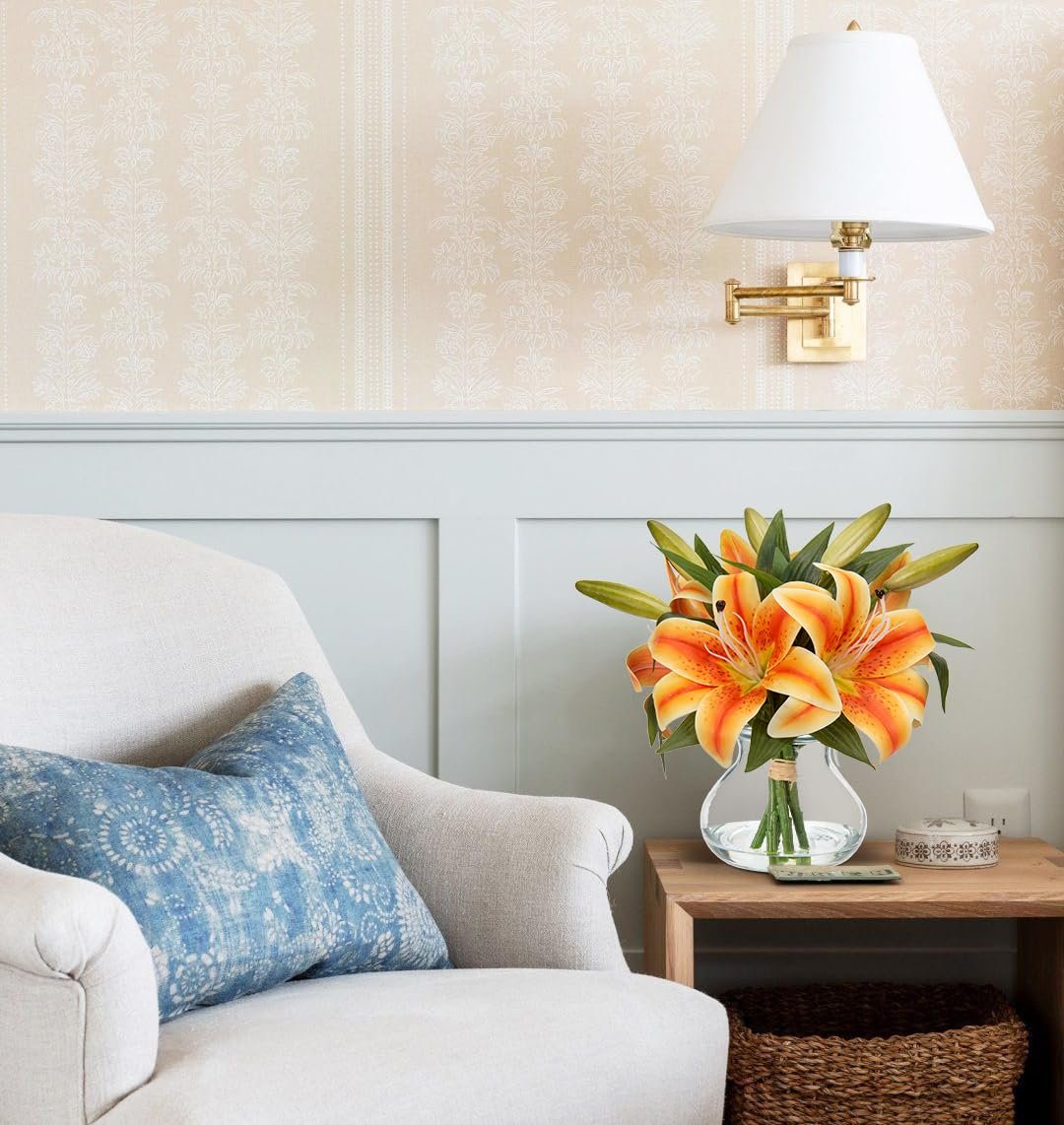 Artificial orange tiger lily arrangement in a glass vase with faux water on a side table next to an armchair.