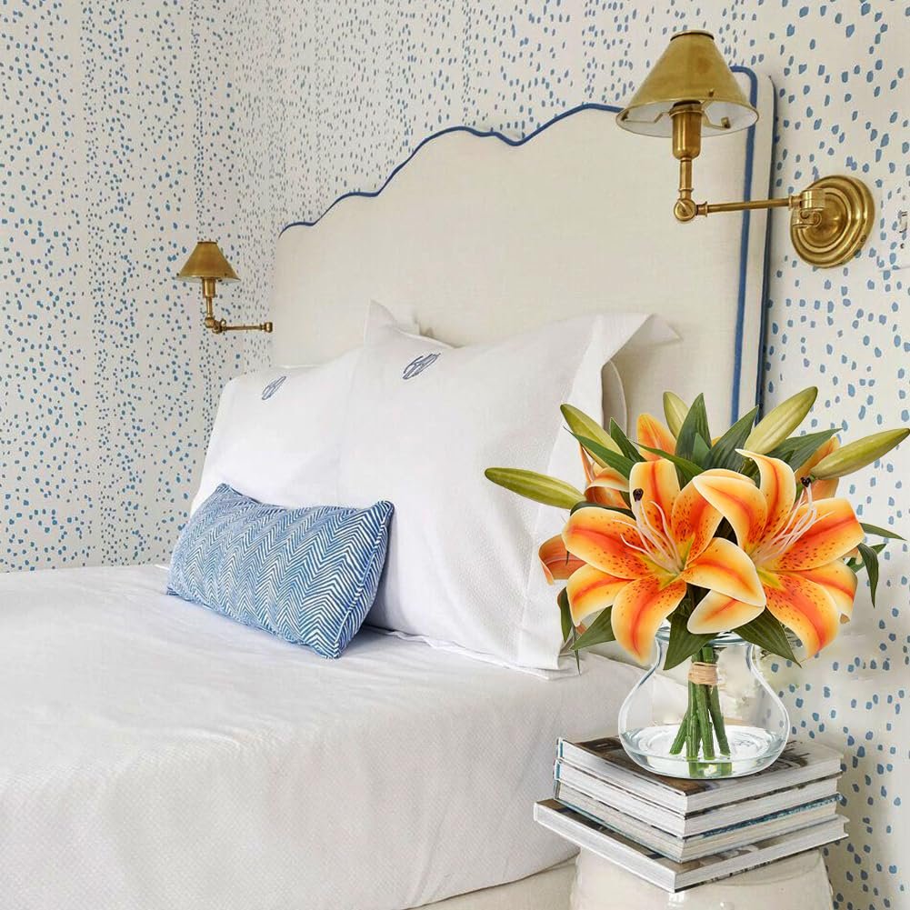 Orange tiger lily artificial flower arrangement in a glass vase on a stack of books next to a bed.