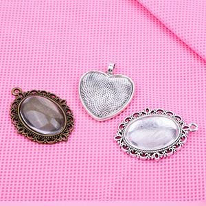 Three metal pendant trays for jewelry making, including oval and heart shapes, on a pink textured surface.