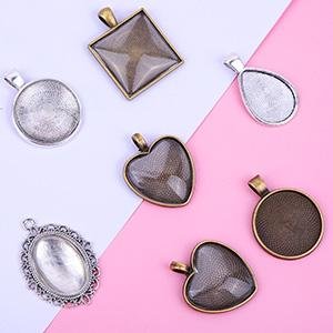 Assortment of six antique-style pendant trays in silver and bronze finishes, including round, oval, heart, and square shapes, arranged on a pink and white background.