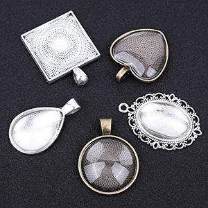 Five assorted metal pendant trays with glass cabochons for jewelry making, including square, heart, teardrop, round, and oval shapes.