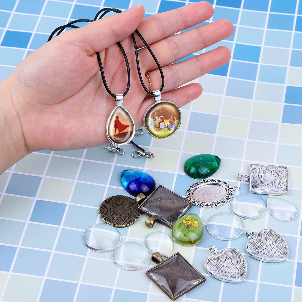 Hand holding two finished pendants with images inside, surrounded by jewelry making supplies like bezels and glass cabochons.