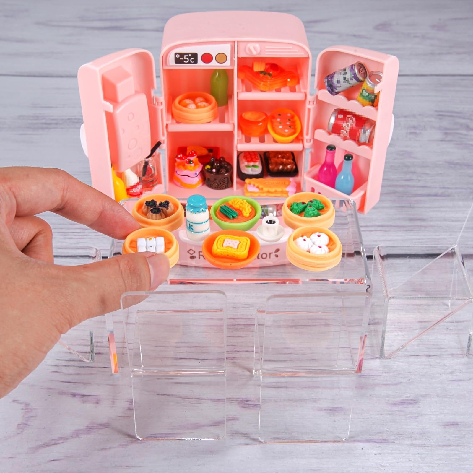 Miniature pink refrigerator filled with tiny food items, next to a table with miniature food dishes and a hand placing a dish.