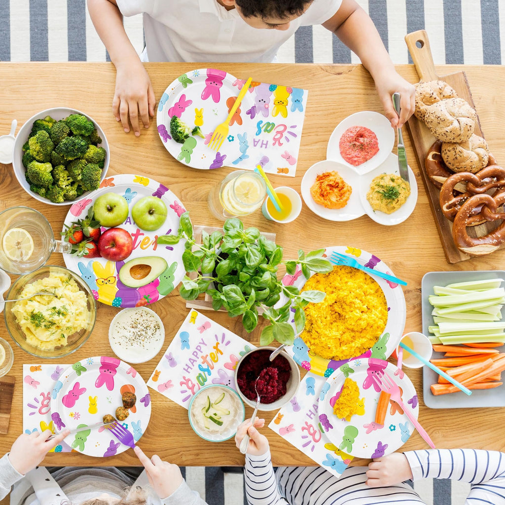 Overhead view of an Easter-themed table setting with plates, napkins, food, and drinks, with children's hands visible.