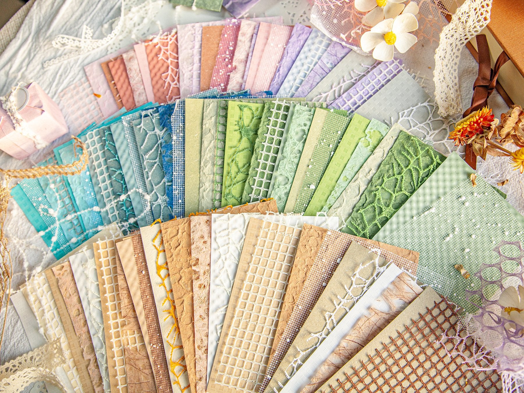 Fan of textured scrapbook paper and mesh fabric sheets in various pastel colors and earth tones, arranged on a white textured surface with decorative elements.