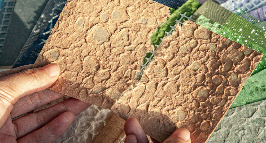 Hands holding a torn piece of textured tan paper over a stack of various textured papers and fabrics in shades of green, blue, and white.