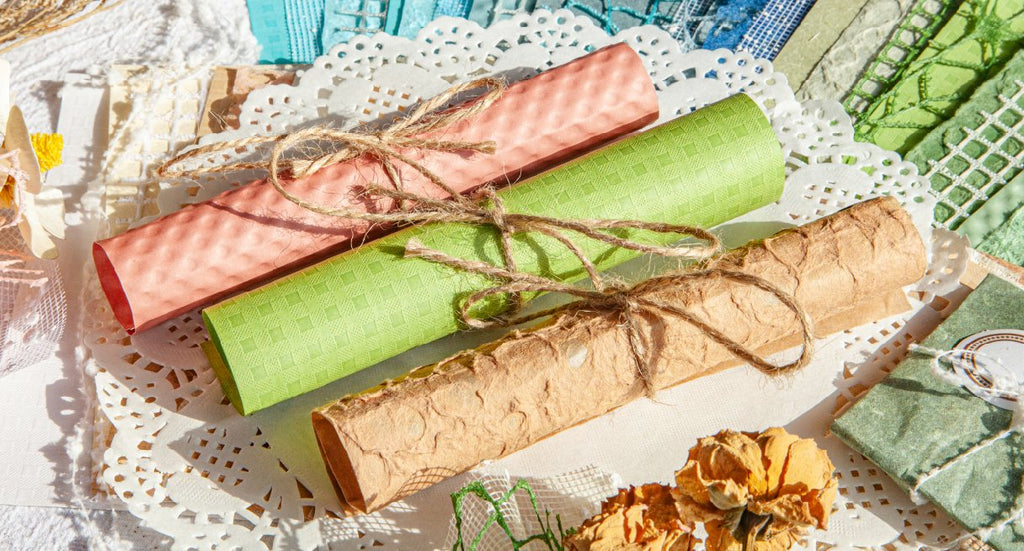 Three rolled textured papers in pink, green, and brown tied with twine, arranged on doilies and craft supplies.