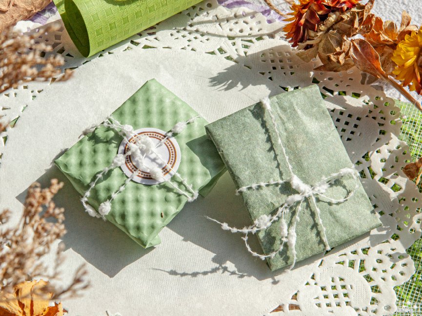 Two green wrapped gifts tied with white string on a white doily, with dried flowers and green textured paper.