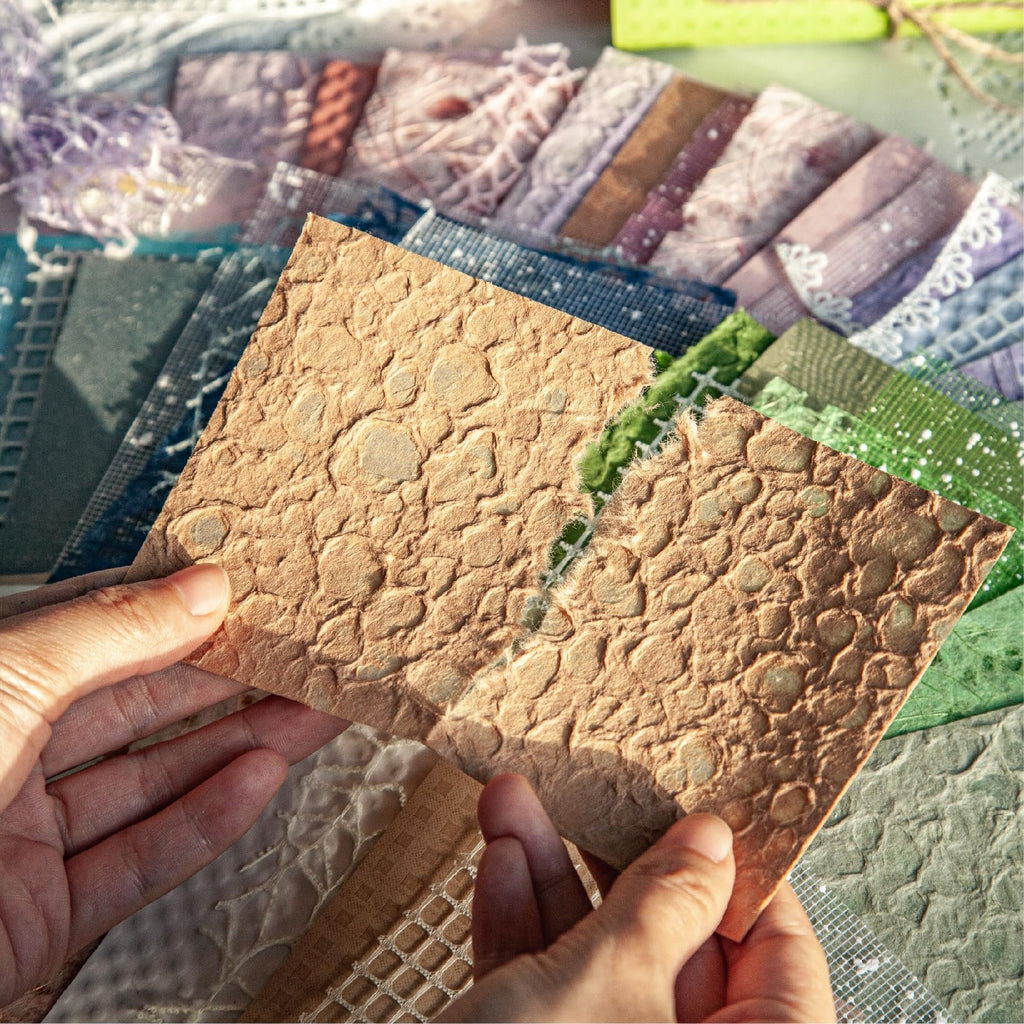 Hands holding textured scrapbook paper with a variety of patterned papers and mesh fabric in the background.