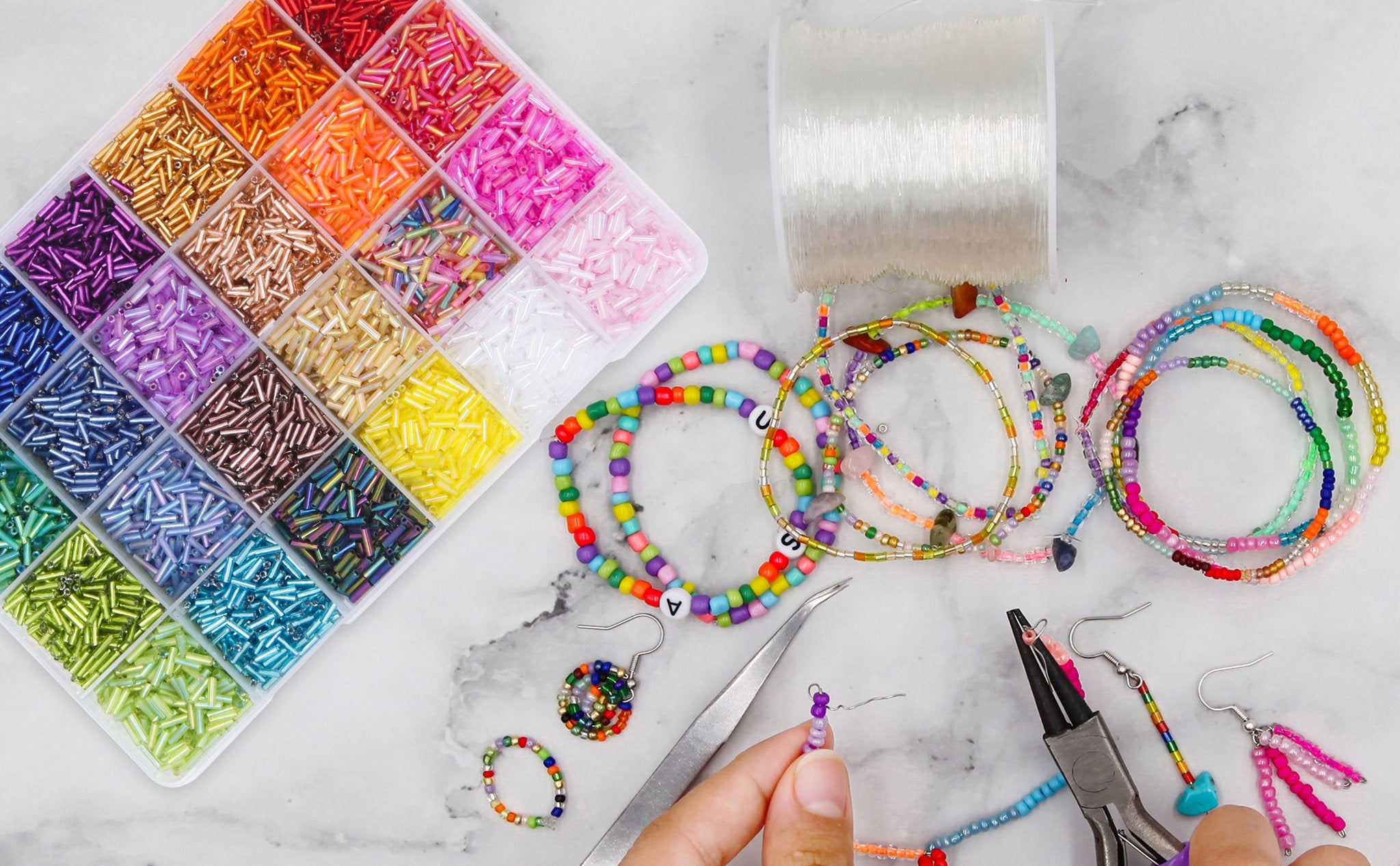 Flat lay of a bead kit with 24 compartments of colorful bugle beads, jewelry making tools, and finished bracelets on a marble surface.
