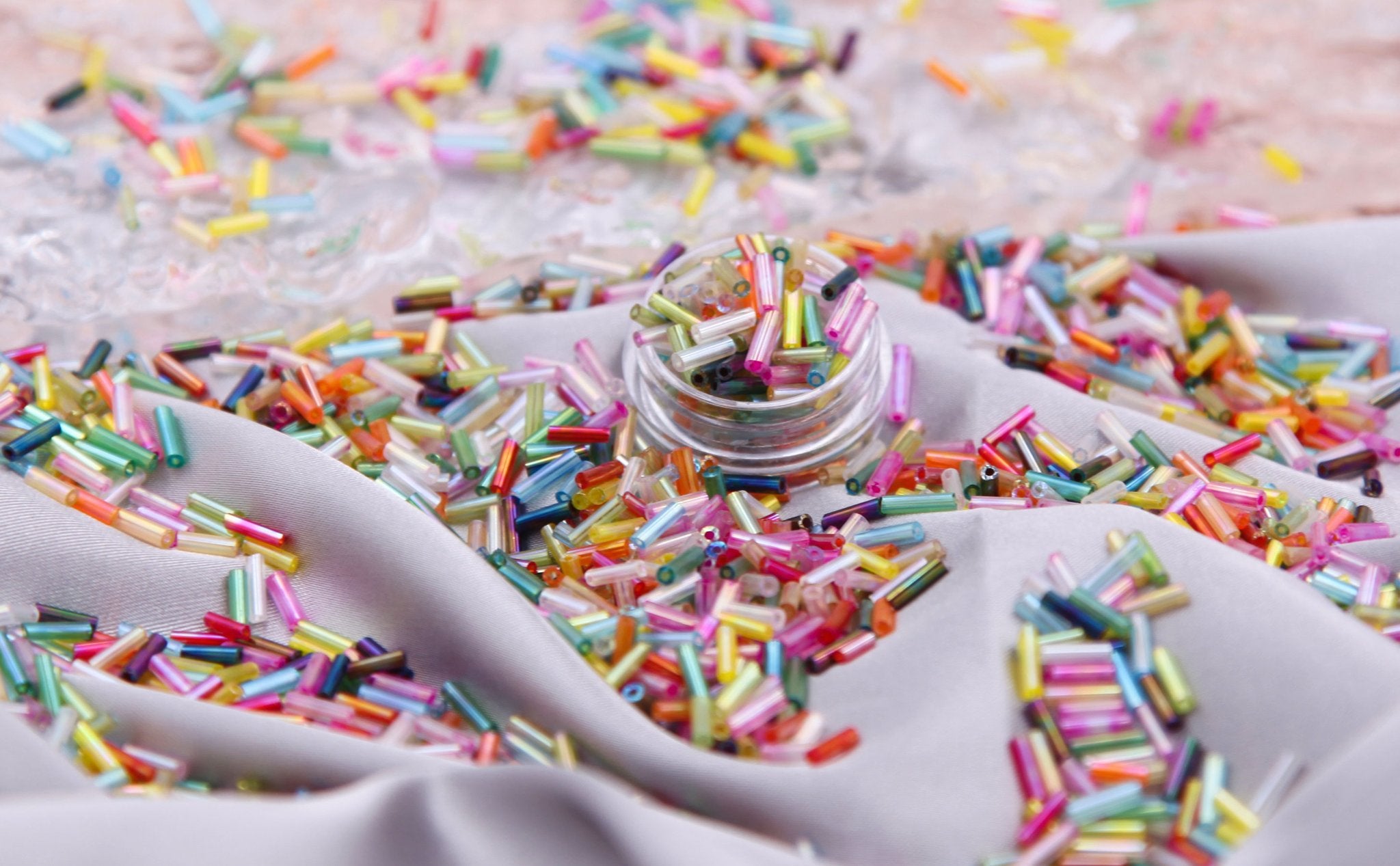 Glass bugle seed beads in a variety of colors spilled on gray fabric with a small clear container of beads.