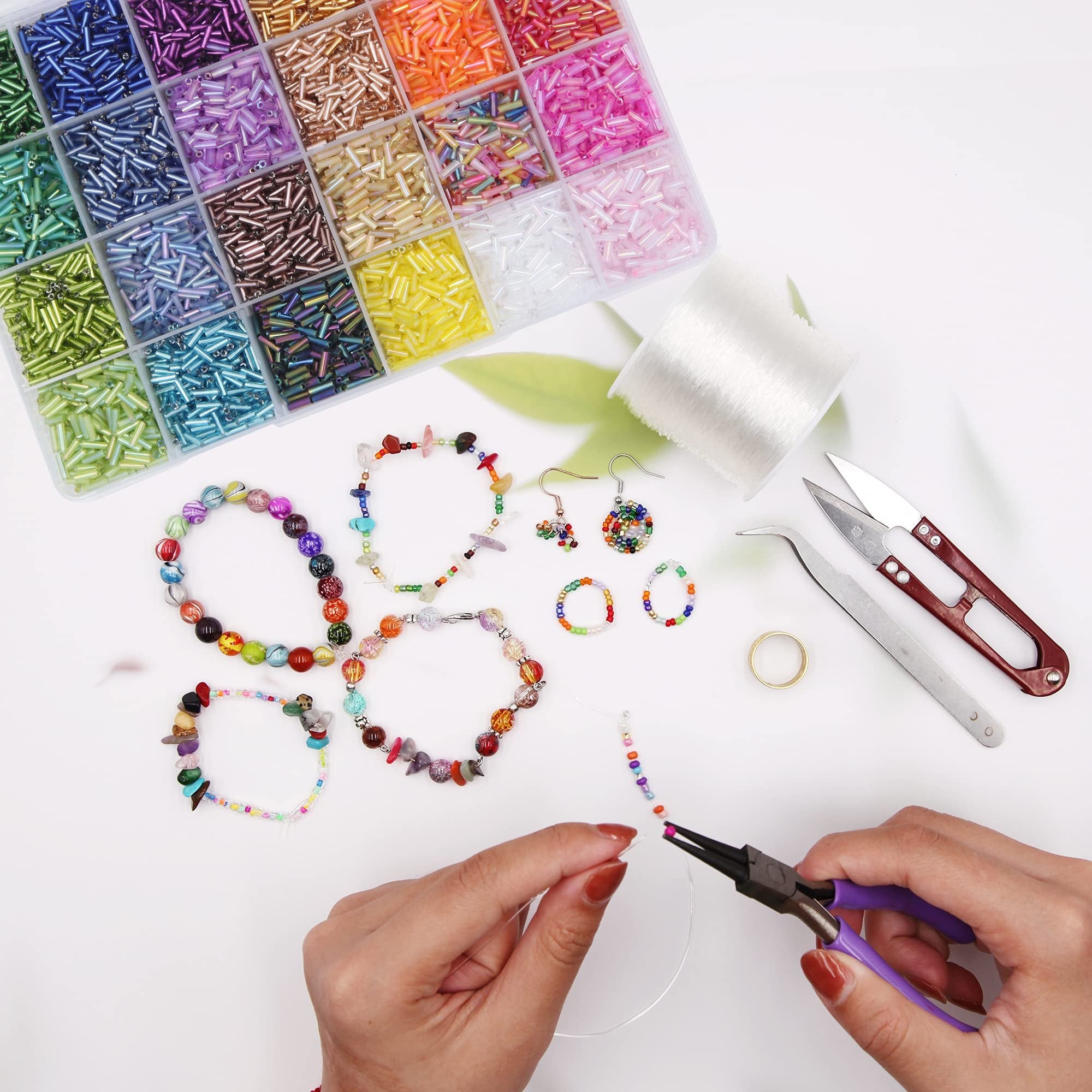 Hands using pliers to string colorful bugle beads onto clear cord, surrounded by finished jewelry and a bead organizer.