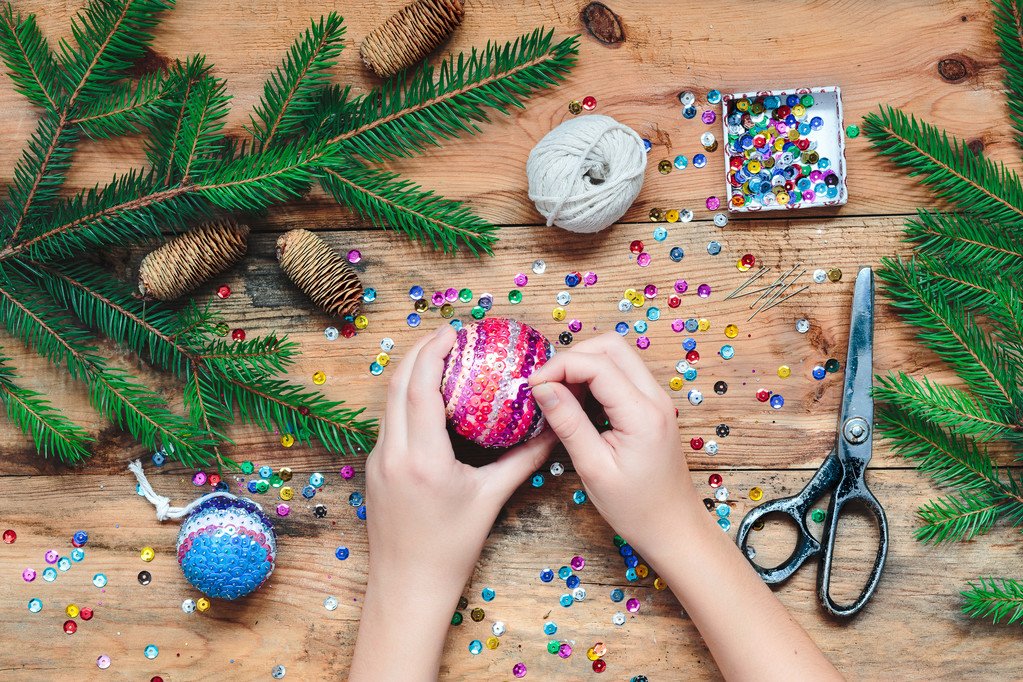 Hands decorating a Christmas ornament with colorful sequins, surrounded by pine branches, pinecones, yarn, and scissors on a wooden table.