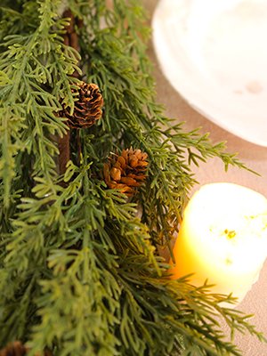 Close-up of a faux evergreen garland with pine cones and a lit candle on a table.