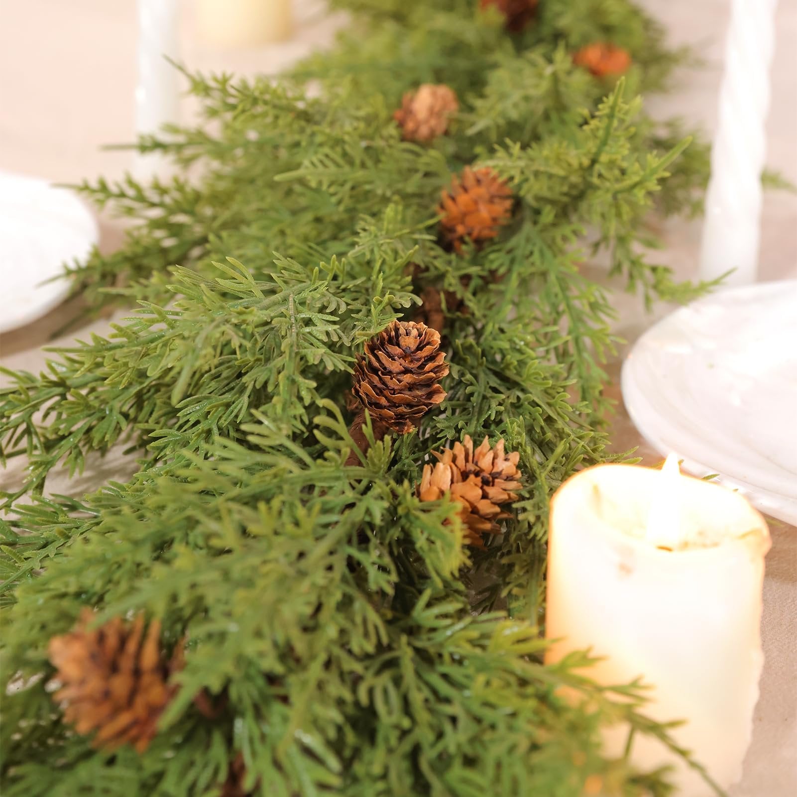 Close-up of a green artificial garland with pine cones, draped on a table with candles and plates.