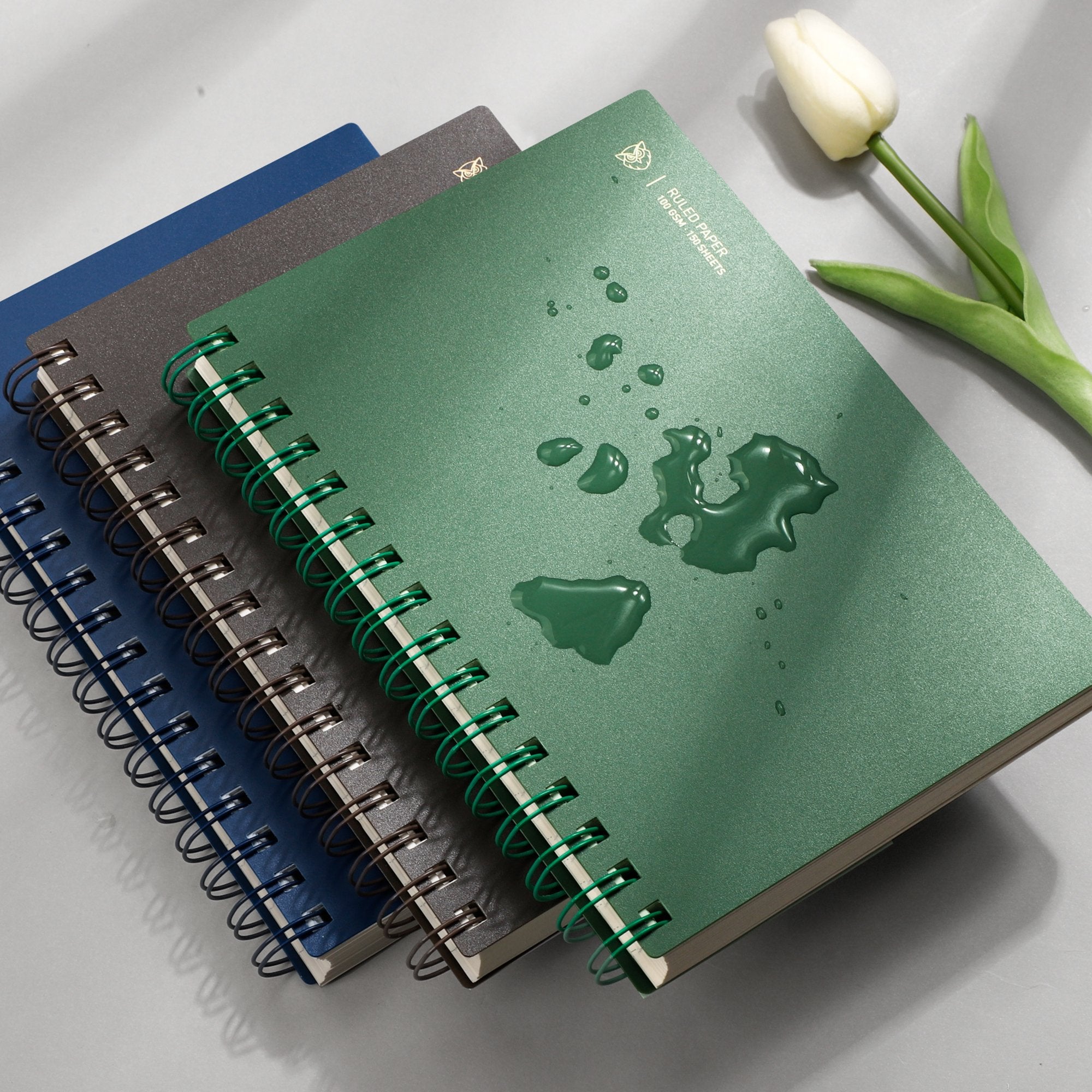Stack of spiral notebooks with a green one in front showing water droplets and a white tulip nearby.