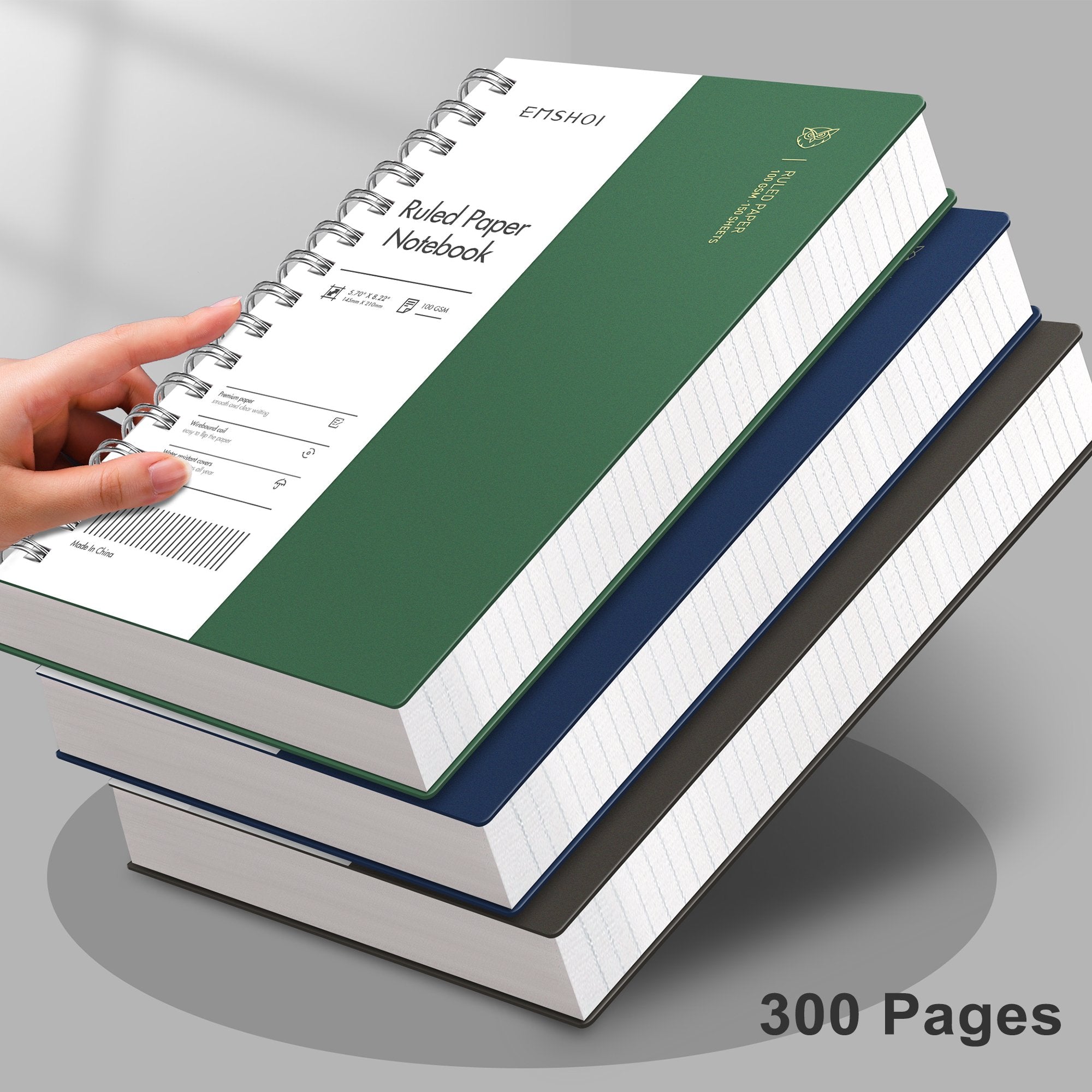 Stack of four spiral notebooks in green, blue, and gray, with a hand holding the top green notebook. Close-up view.