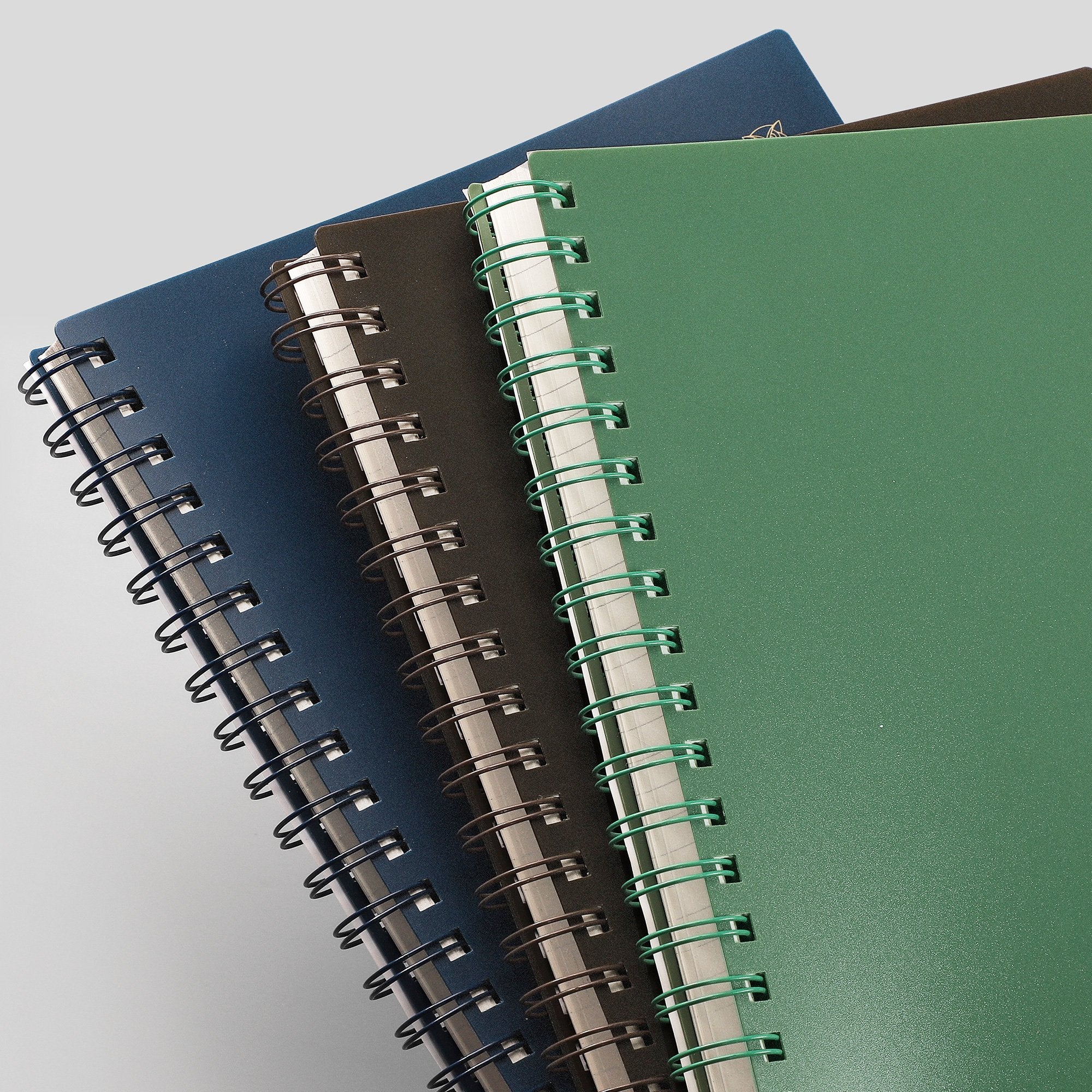 Three spiral-bound notebooks in green, dark blue, and black, angled on a light gray background.