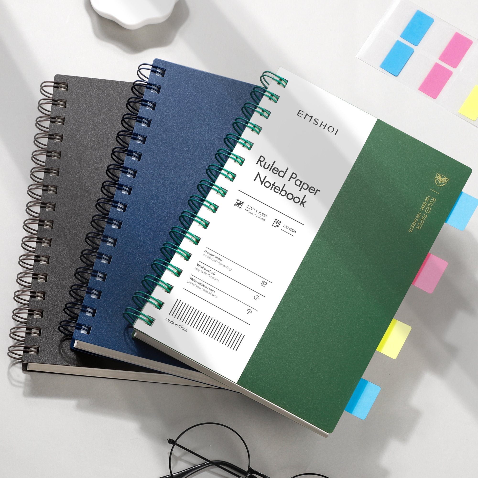 Three spiral notebooks stacked, one green, one blue, one black, with colorful sticky tabs protruding.