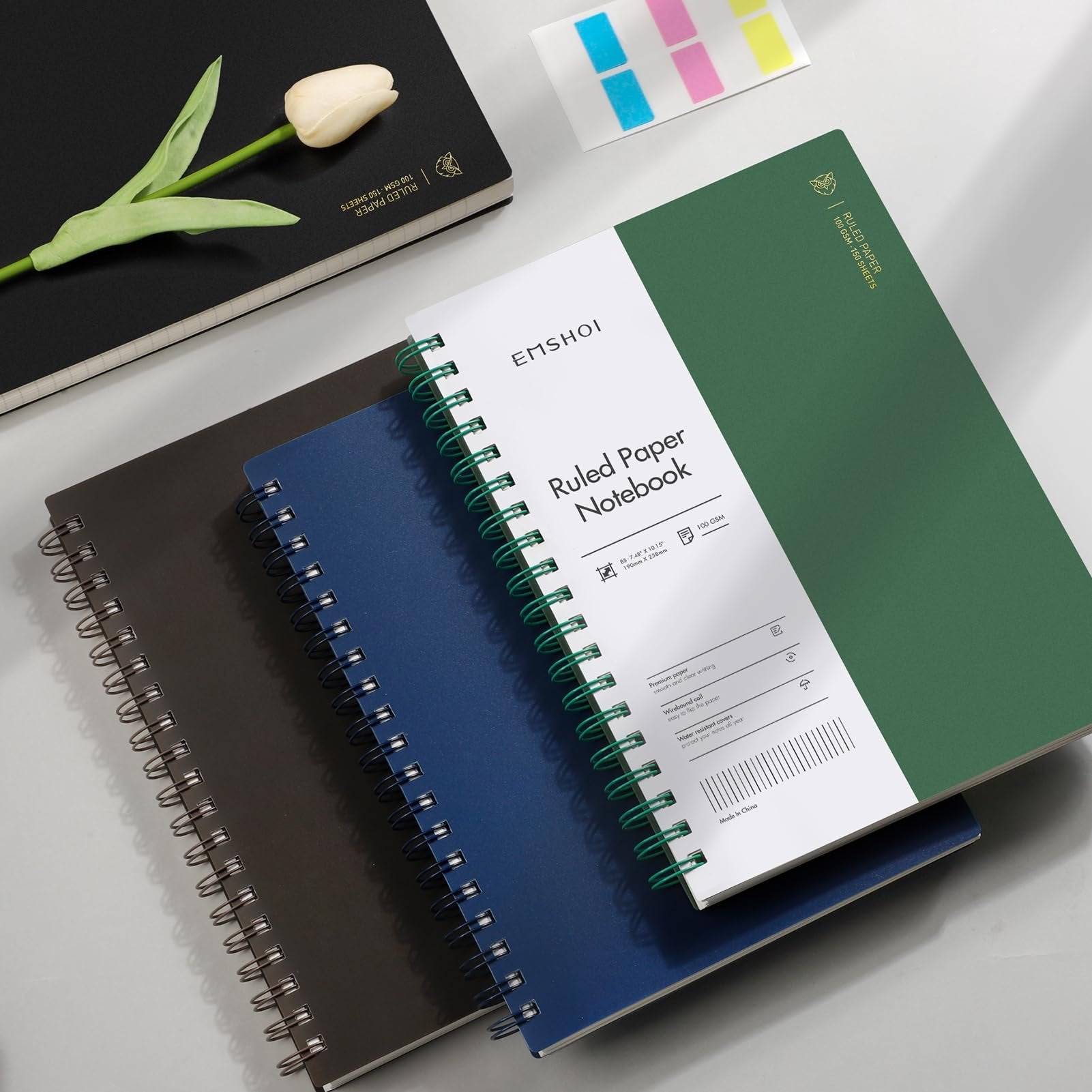 Three spiral-bound notebooks in black, blue, and green, with a white page showing product details and a tulip on the black notebook.