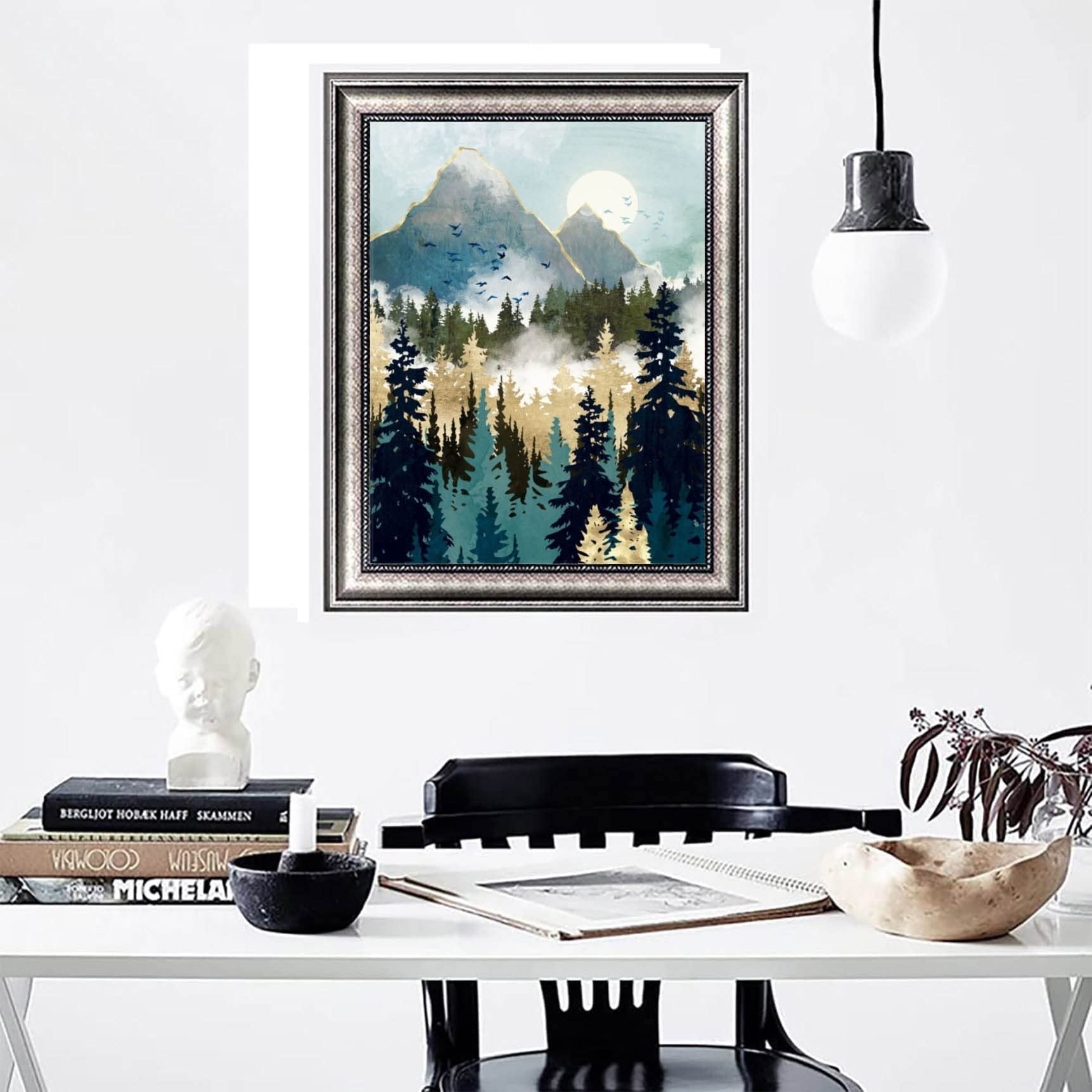 Framed abstract mountain forest landscape painting hanging on a white wall above a desk with books and a bust.