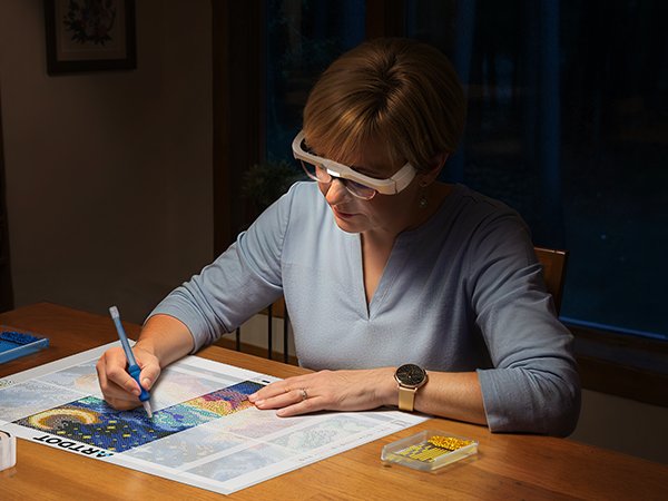 Woman wearing magnifying glasses doing diamond painting at a wooden table, holding a diamond pen