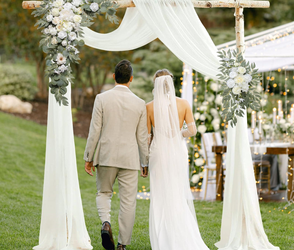 Wedding couple walking through an arch decorated with white flowers, greenery, and draped fabric.
