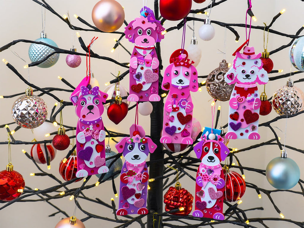 Pink dog-shaped craft ornaments with glitter and gems hanging on a lighted tree branch with Christmas ornaments.