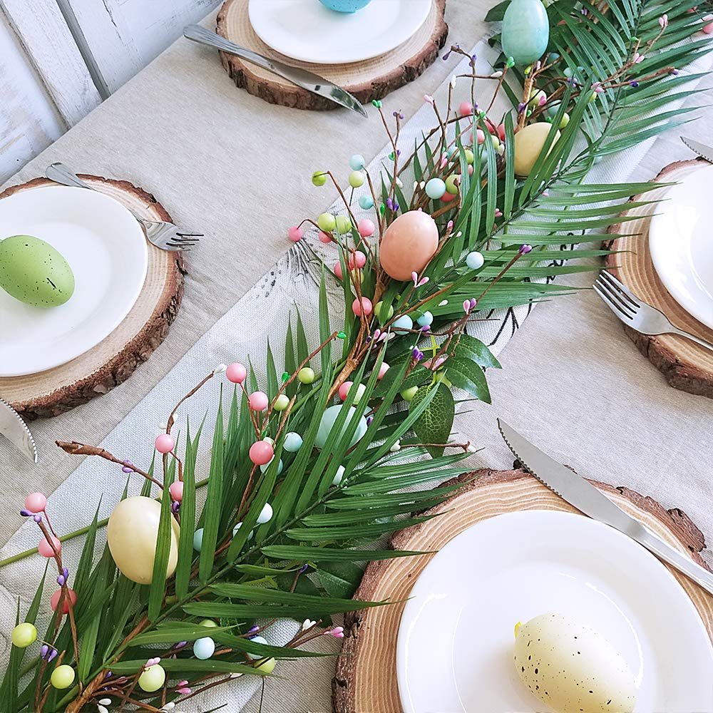 Easter table setting with a garland of artificial palm leaves, colorful berries, and decorative eggs on a runner.