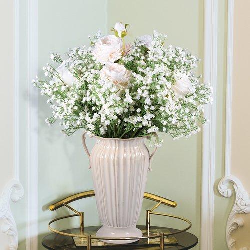 White artificial baby's breath flowers and roses in a cream-colored vase on a table.