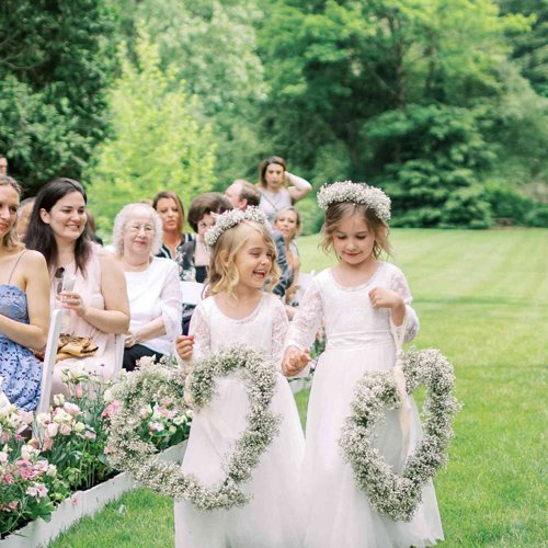 Two young flower girls in white dresses walk down an aisle holding heart-shaped wreaths of baby's breath flowers.