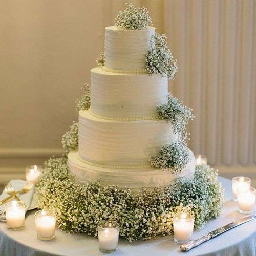 Four-tier white frosted wedding cake decorated with white baby's breath flowers and surrounded by more flowers and candles.