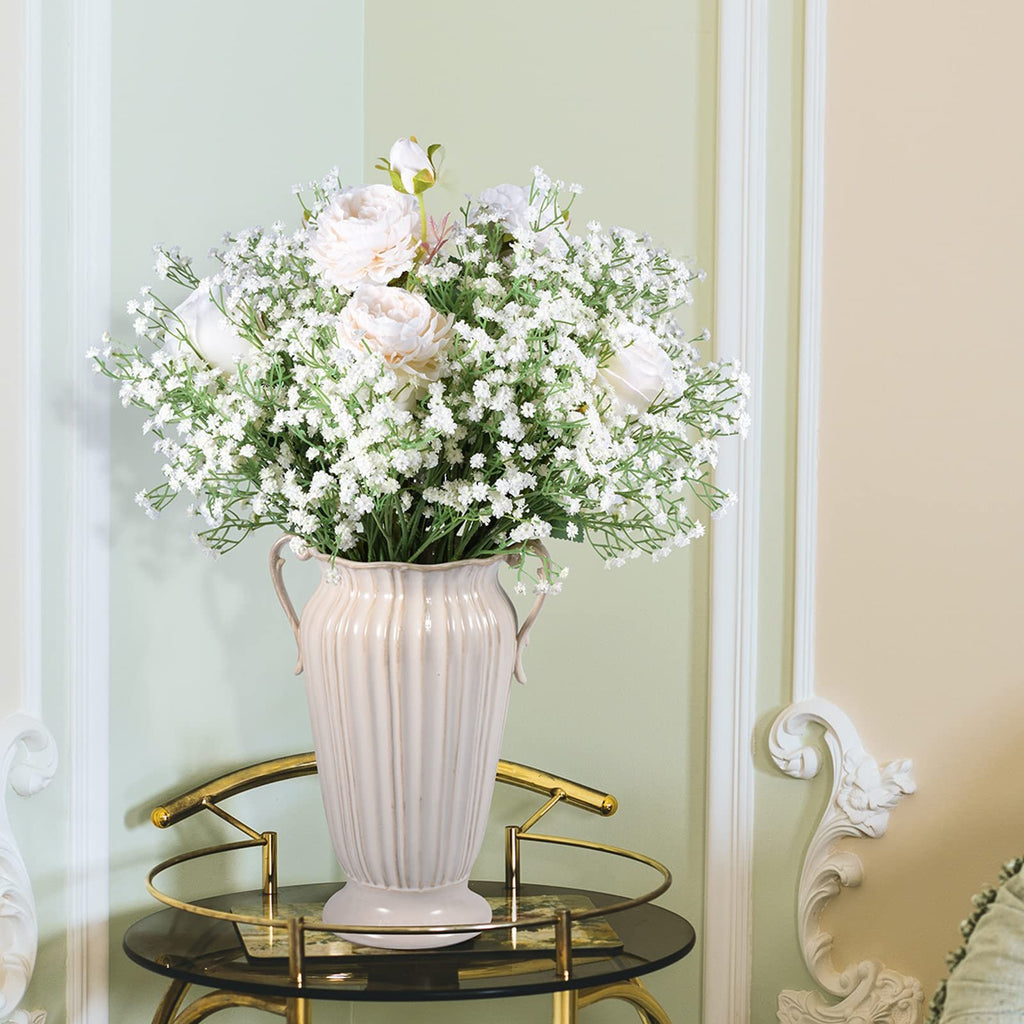 White artificial baby's breath and peony flowers in a cream vase on a glass table.