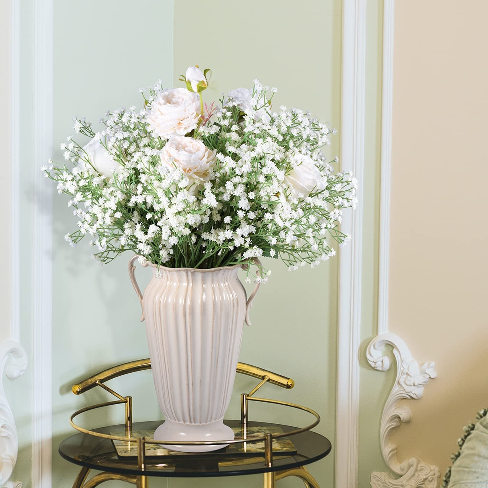 White artificial baby's breath and peony flowers in a cream vase on a glass table.