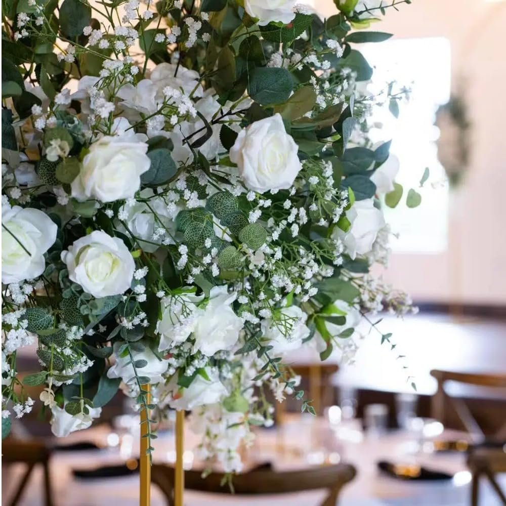 Close-up of a white artificial baby's breath and rose floral arrangement on a gold stand, set against a blurred background of a table setting.