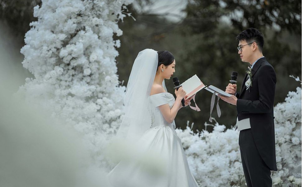 Bride and groom at wedding ceremony, both holding microphones and reading from books, surrounded by white floral decorations.