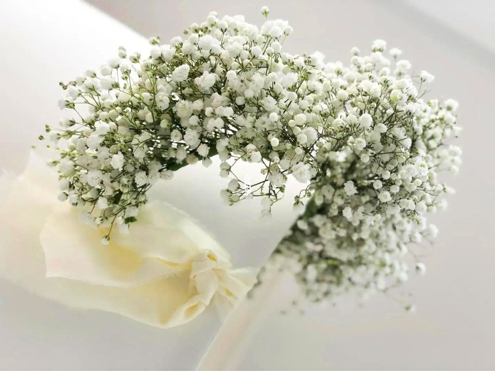 White artificial baby's breath flower crown with a cream ribbon tied around it, displayed on a white surface.