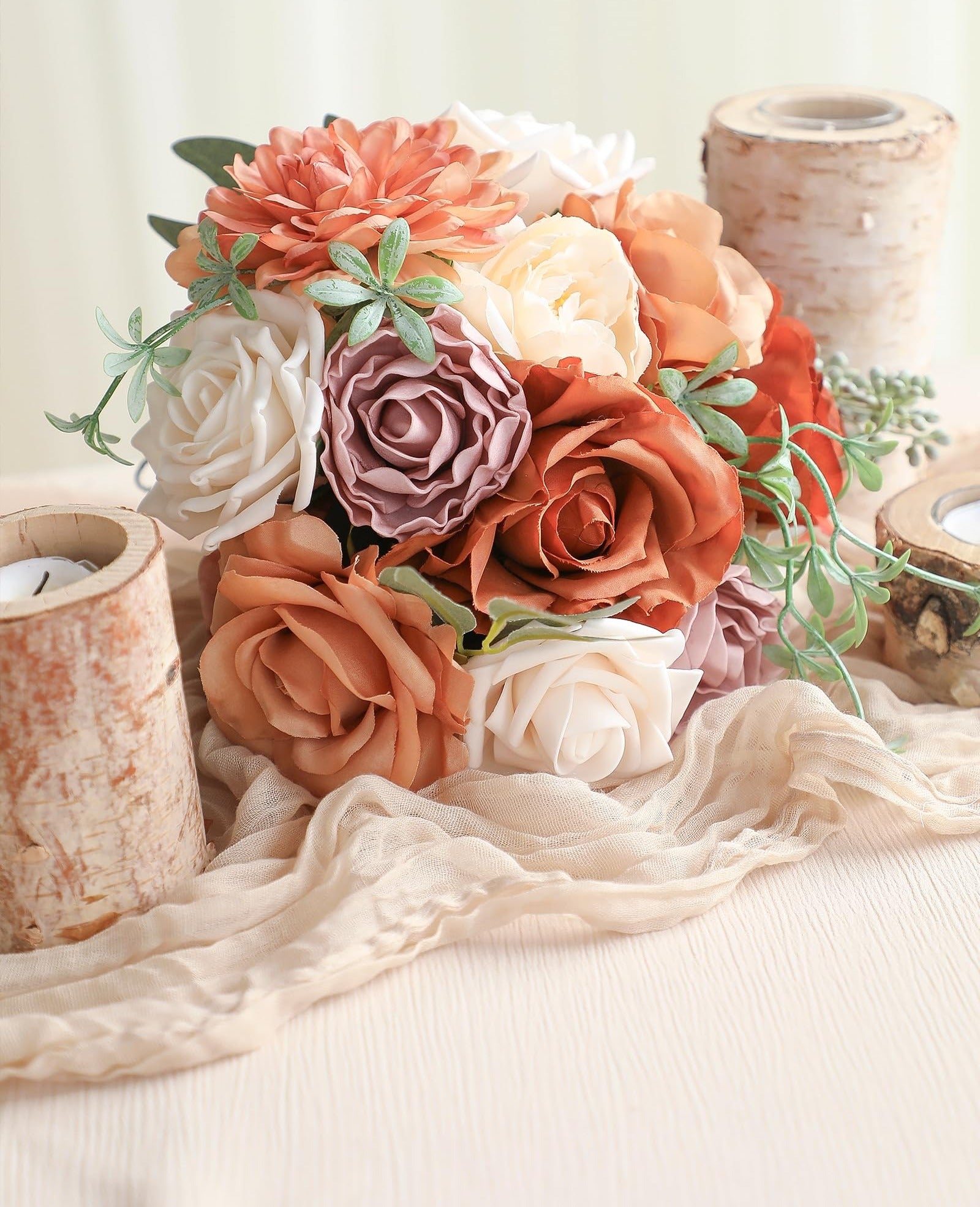 Artificial flower bouquet with terracotta dahlias, champagne peonies, amber roses, and cream roses, arranged with greenery and displayed with birch candle holders and beige fabric.