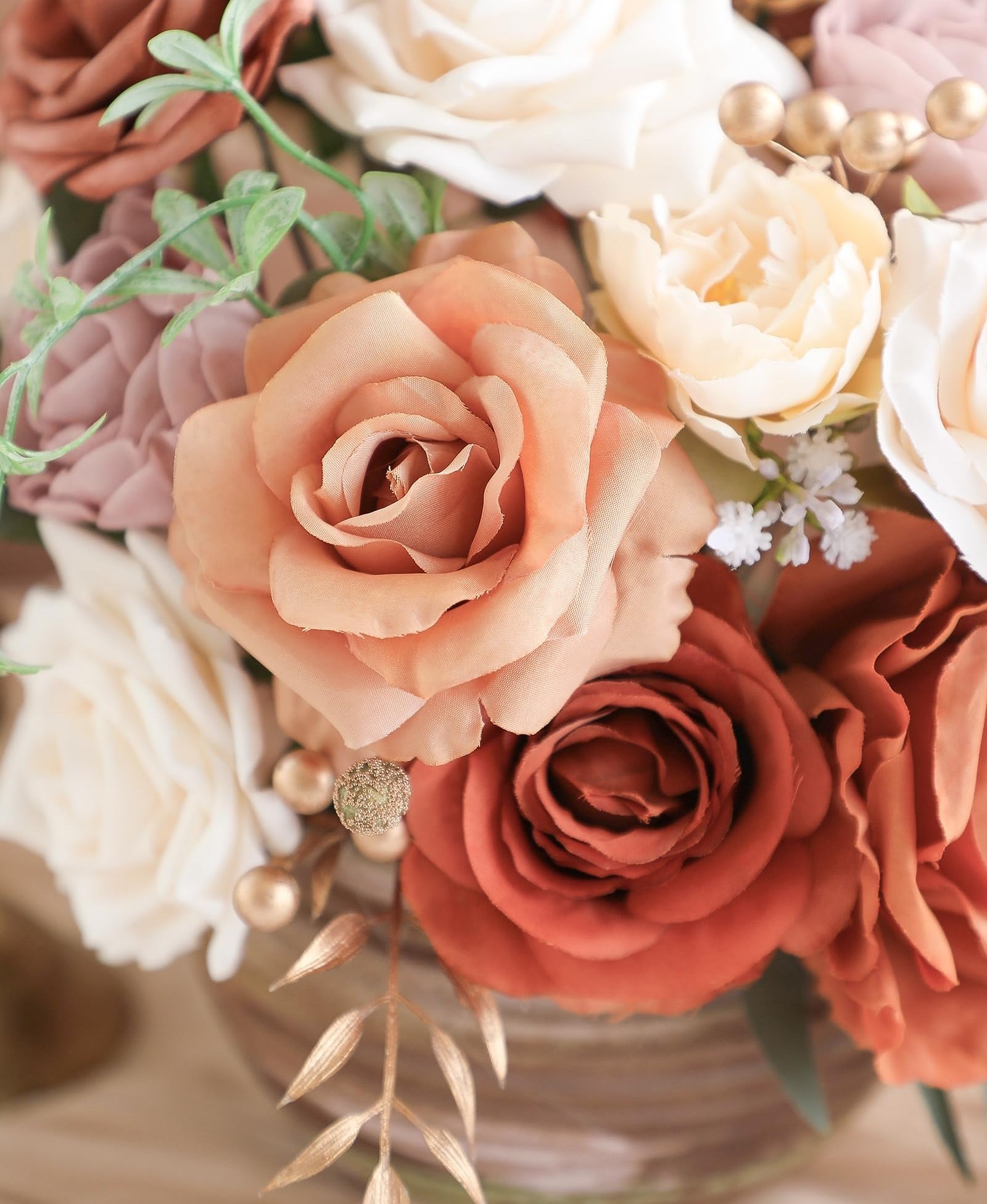 Close-up of an artificial flower arrangement with terracotta, cream, and burnt orange roses, accented with gold berries and green leaves.