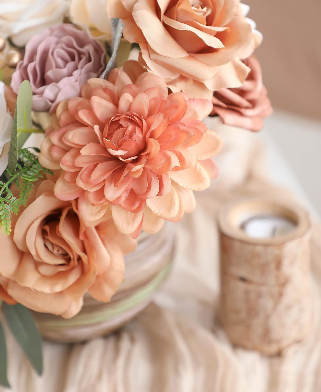 Close-up of a floral arrangement with terracotta dahlias, champagne roses, and dusty rose peonies in a vase, with a wooden candle holder to the right.