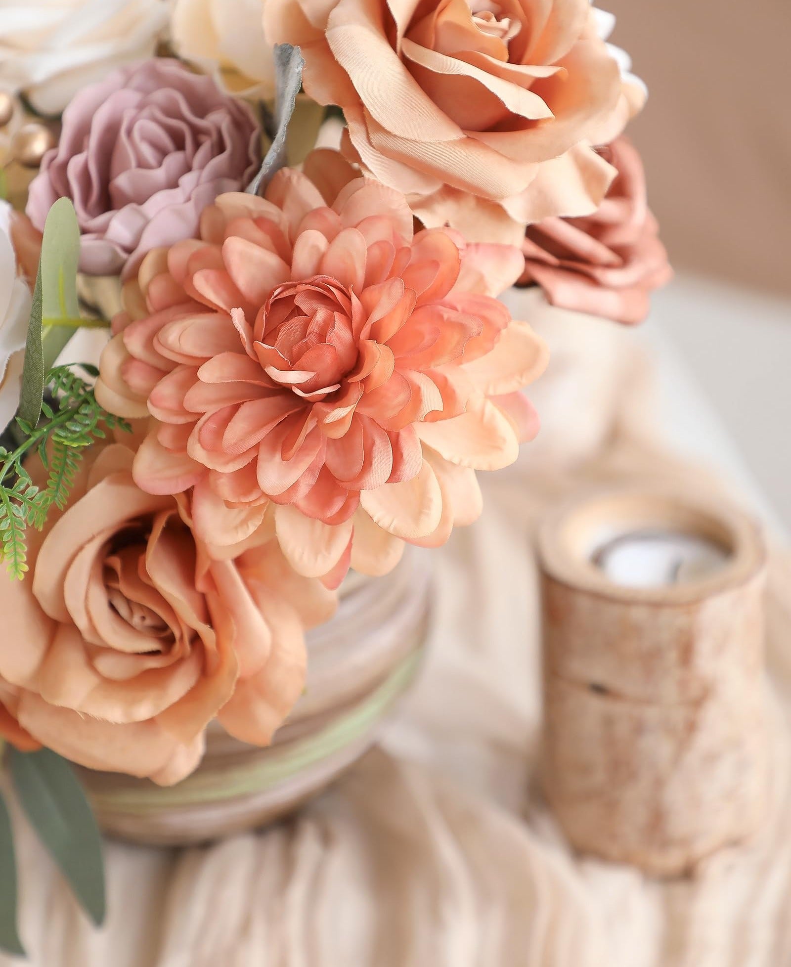 Close-up of a floral arrangement with terracotta dahlias, champagne roses, and dusty rose peonies in a vase, with a wooden candle holder to the right.