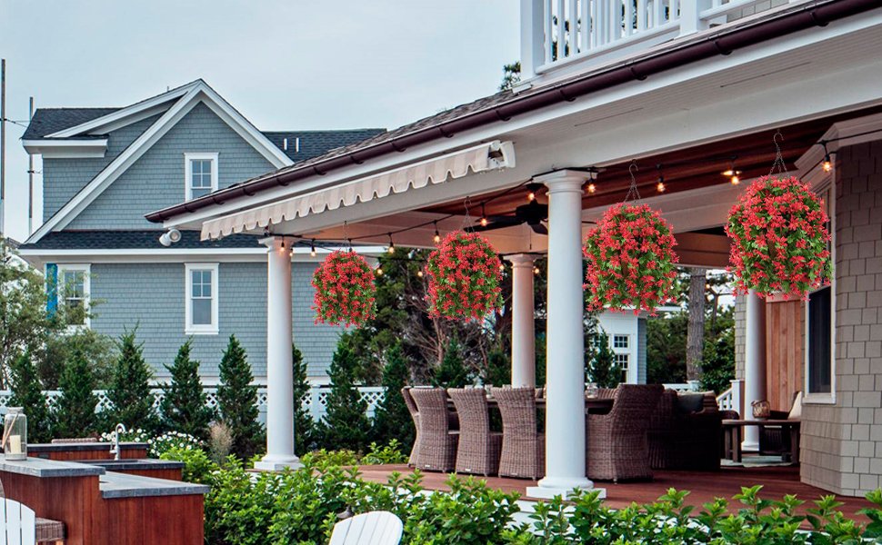Four hanging baskets with red flowers and green leaves suspended from a porch ceiling, decorating an outdoor patio area with furniture.