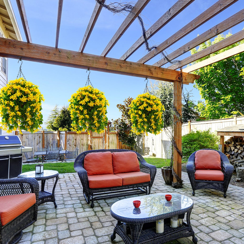 Three hanging baskets filled with yellow artificial flowers suspended from a wooden pergola over a patio with outdoor furniture.