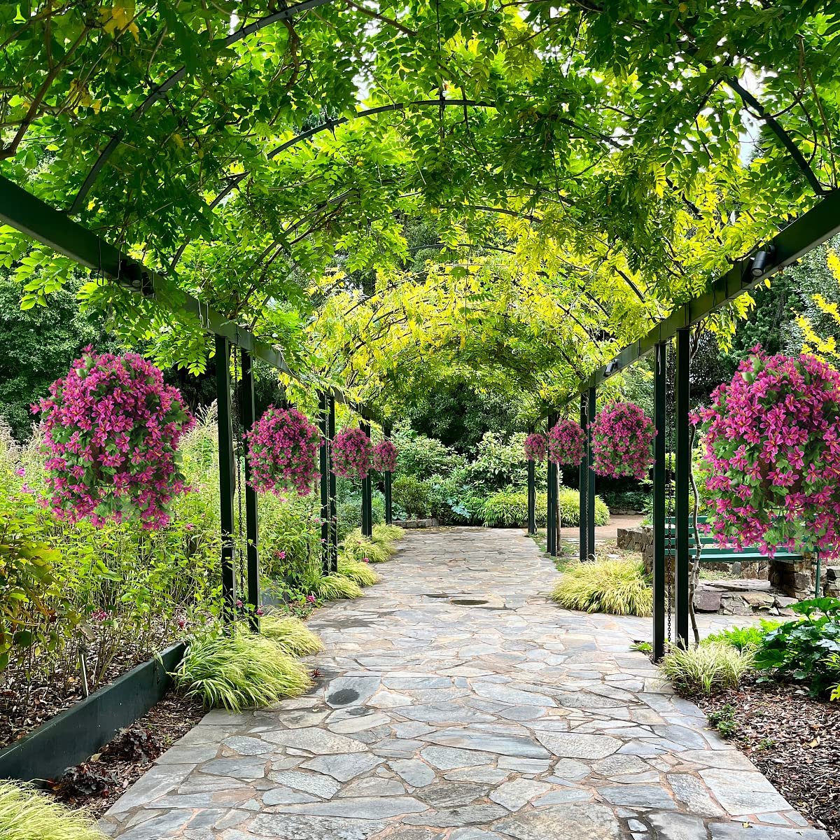 Stone pathway through a garden arbor covered in green leaves with hanging baskets of pink flowers