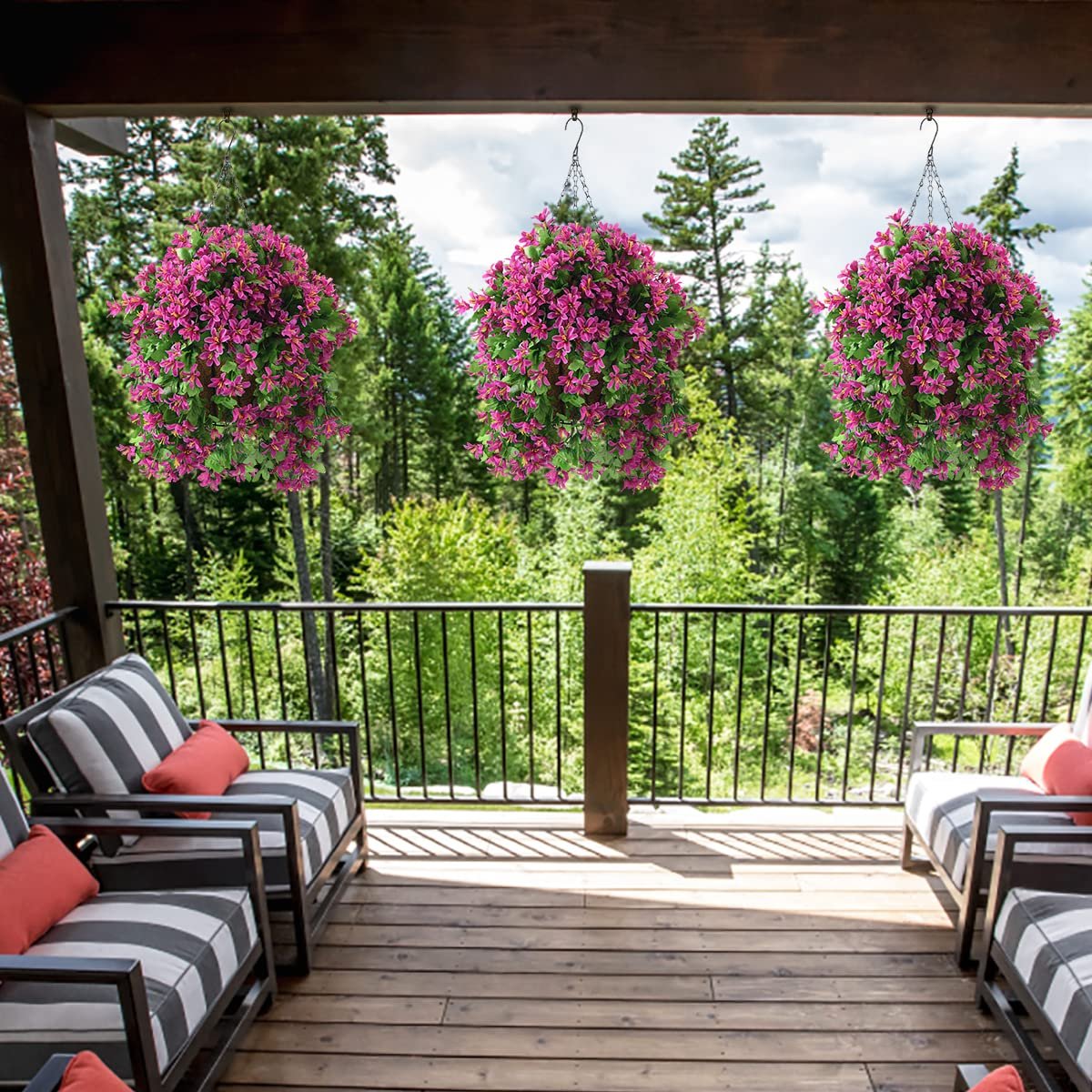 Three hanging baskets of artificial pink violet flowers with green leaves on a wooden deck with chairs and a forest background.