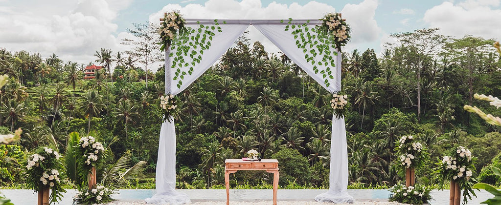 Wedding arch decorated with white fabric and artificial ivy vines, set against a lush tropical forest backdrop.